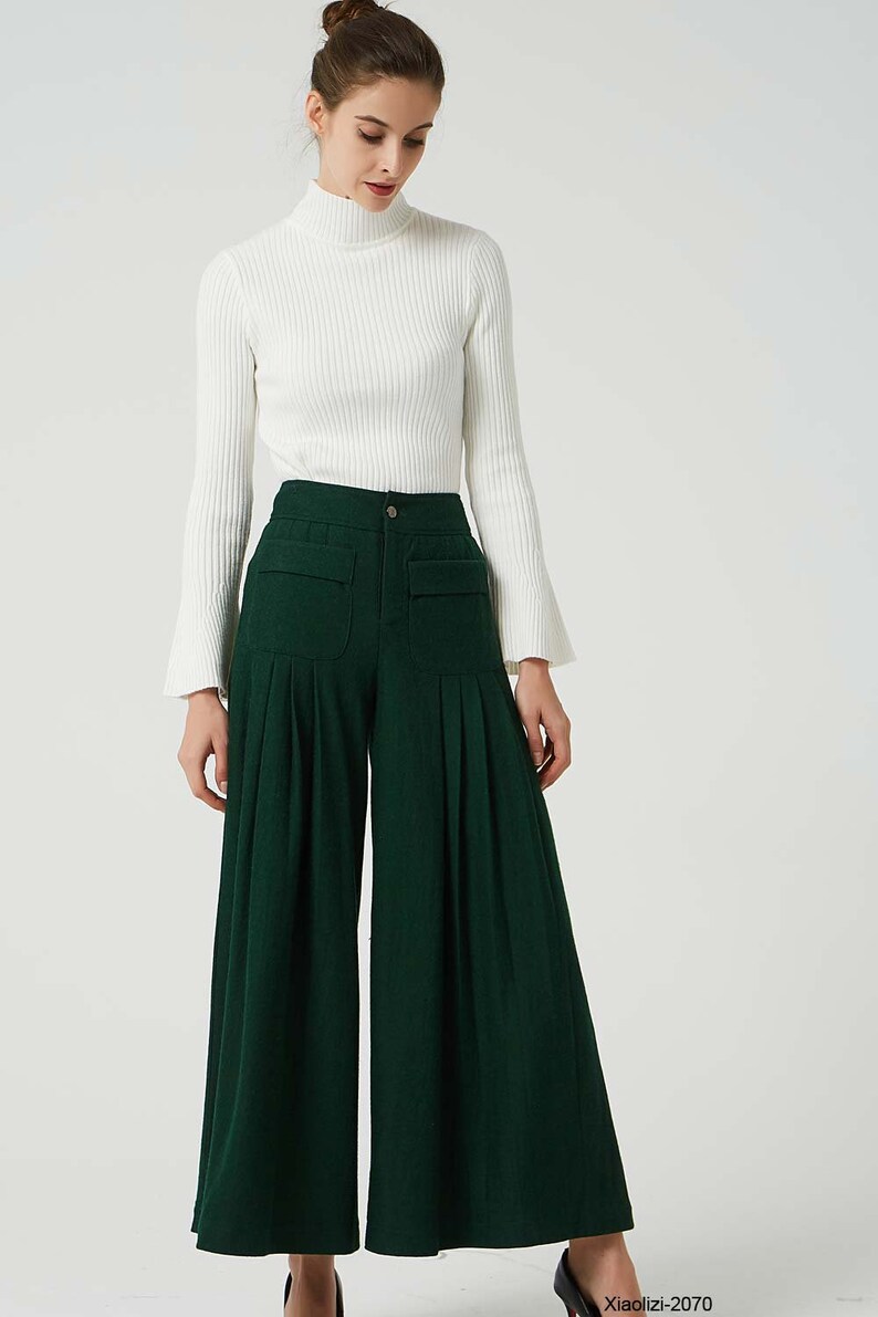 May include: A woman is wearing a white turtleneck sweater and dark green wide-leg trousers with pleats. The trousers have large pockets on the front.