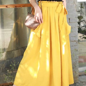 May include: A woman wearing a black top and a long, flowing yellow skirt with a tied waist and pockets. She is also wearing black sandals and carrying a small woven purse. The skirt is labeled "6-yellow".