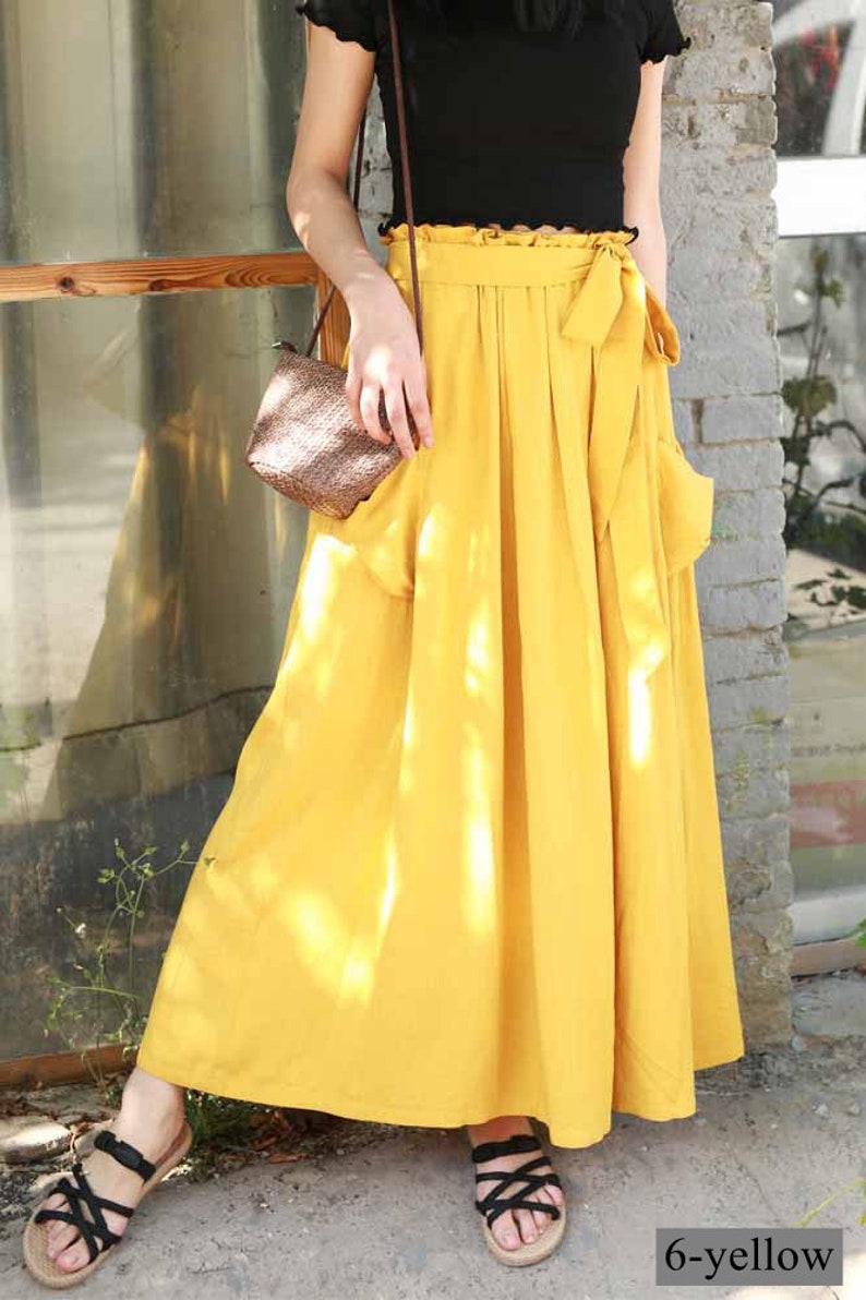 May include: A woman wearing a black top and a long, flowing yellow skirt with a tied waist and pockets. She is also wearing black sandals and carrying a small woven purse. The skirt is labeled "6-yellow".