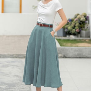 May include: A woman wearing a white t-shirt, a brown belt, a light blue midi skirt, and black heels. She is wearing a straw hat with a white band.