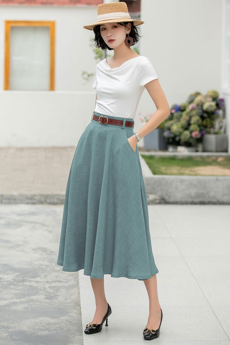 May include: A woman wearing a white t-shirt, a brown belt, a light blue midi skirt, and black heels. She is wearing a straw hat with a white band.