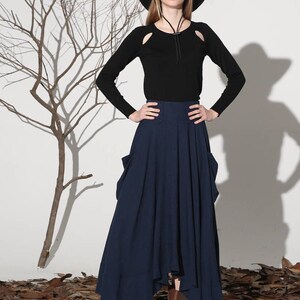 Op de afbeelding: Een vrouw draagt een zwart shirt met lange mouwen met uitsnijdingen op de schouders, een brede zwarte hoed, een donkerblauwe maxi-rok met zakken en bruine enkellaarsjes.