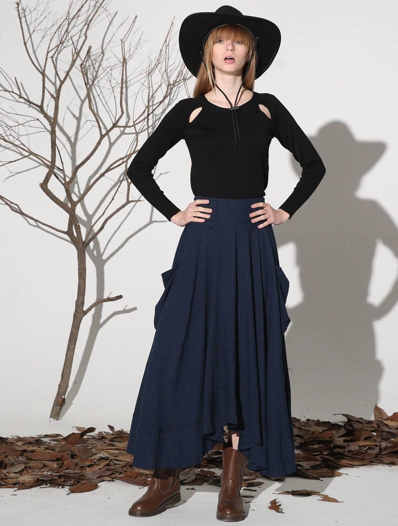 Op de afbeelding: Een vrouw draagt een zwart shirt met lange mouwen met uitsnijdingen op de schouders, een brede zwarte hoed, een donkerblauwe maxi-rok met zakken en bruine enkellaarsjes.