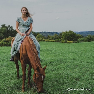 Op de afbeelding: Een vrouw in een bloemenjurk rijdt op een bruin paard in een grasveld. Het paard graast op het gras. De vrouw glimlacht en kijkt naar de camera. De achtergrond is een groen veld met bomen in de verte.