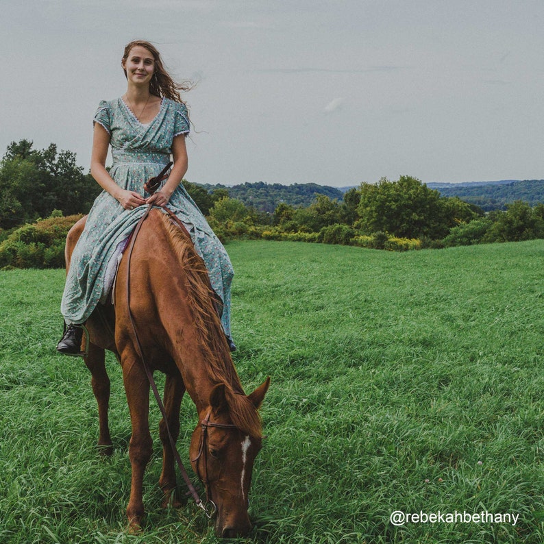 Op de afbeelding: Een vrouw in een bloemenjurk rijdt op een bruin paard in een grasveld. Het paard graast op het gras. De vrouw glimlacht en kijkt naar de camera. De achtergrond is een groen veld met bomen in de verte.