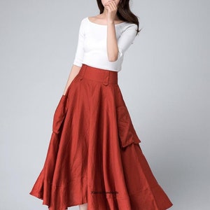 Op de afbeelding: Een roestkleurige, hoge taille, linnen rok met twee grote zakken aan de voorkant. De rok is A-lijn en valt tot aan de enkels. De rok wordt gedragen met een wit, kort mouwig shirt.