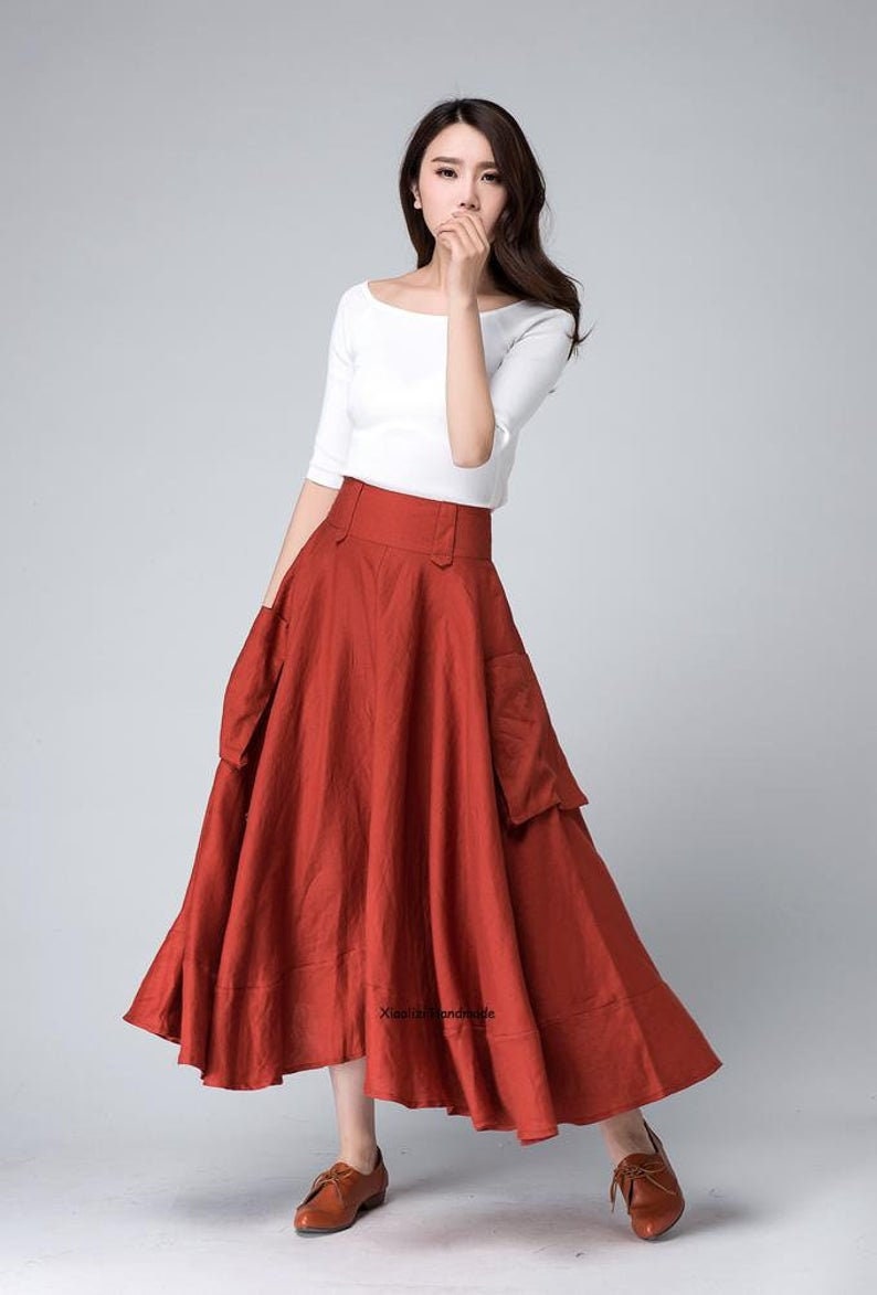 Op de afbeelding: Een roestkleurige, hoge taille, linnen rok met twee grote zakken aan de voorkant. De rok is A-lijn en valt tot aan de enkels. De rok wordt gedragen met een wit, kort mouwig shirt.