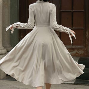 May include: A beige long-sleeved dress with a cinched waist and a flowing skirt. The dress has a collar and long sleeves with ties at the cuffs.