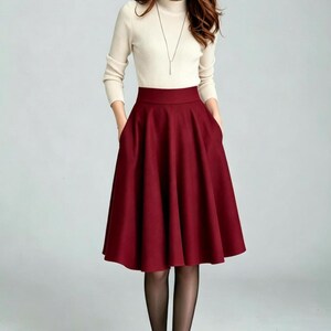 May include: A cream-colored long-sleeve mock neck sweater paired with a high-waisted, knee-length, deep burgundy A-line skirt featuring side pockets. The outfit is completed with sheer black tights and dark-colored closed-toe pumps.