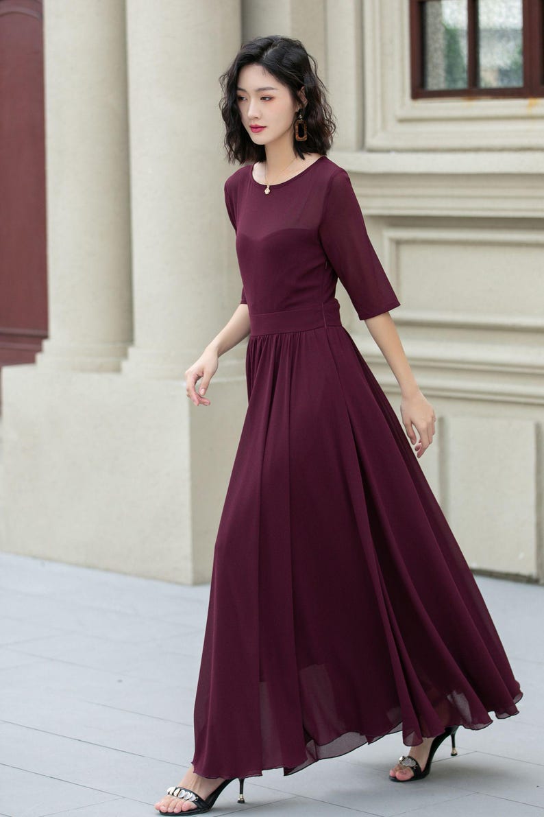May include: A woman wearing a long, burgundy maxi dress with a round neckline and short sleeves. The dress has a fitted bodice and a flowing skirt. She is wearing black heeled sandals.