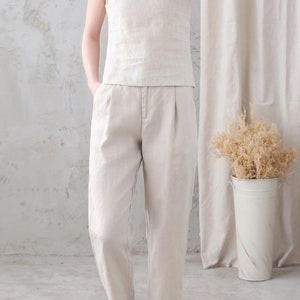Pu&ograve; includere: Una donna indossa una canotta di lino beige chiaro senza maniche e pantaloni di lino abbinati. Indossa sandali bianchi.