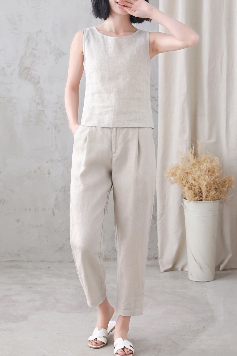 Pu&ograve; includere: Una donna indossa una canotta di lino beige chiaro senza maniche e pantaloni di lino abbinati. Indossa sandali bianchi.