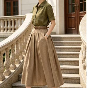 May include: An olive green button-down shirt paired with a high-waisted, pleated, midi-length skirt in a tan color. A brown belt cinches the waist. The outfit is accessorized with a brown hat and black heeled sandals. The ensemble is set against a backdrop of stone steps and a building.