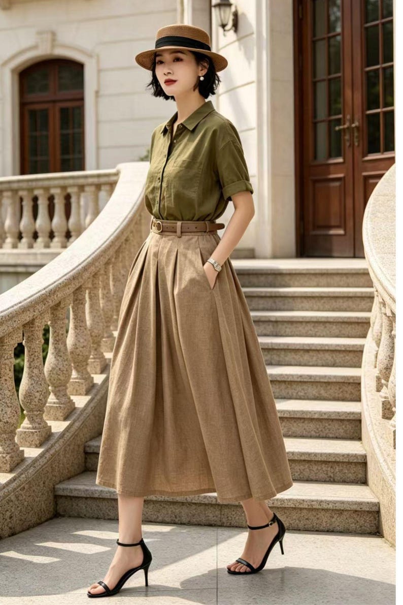 May include: An olive green button-down shirt paired with a high-waisted, pleated, midi-length skirt in a tan color. A brown belt cinches the waist. The outfit is accessorized with a brown hat and black heeled sandals. The ensemble is set against a backdrop of stone steps and a building.