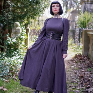 May include: A woman wearing a long, dark purple dress with a black leather harness and belt. She is standing in a cemetery with green grass and fallen leaves.