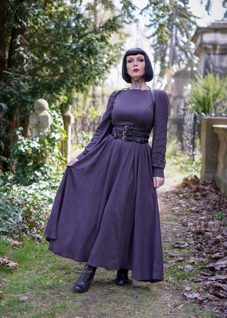 May include: A woman wearing a long, dark purple dress with a black leather harness and belt. She is standing in a cemetery with green grass and fallen leaves.