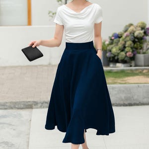 May include: A woman wearing a white short-sleeved top and a dark blue midi skirt with a high waist. She is wearing black heels with gold accents.