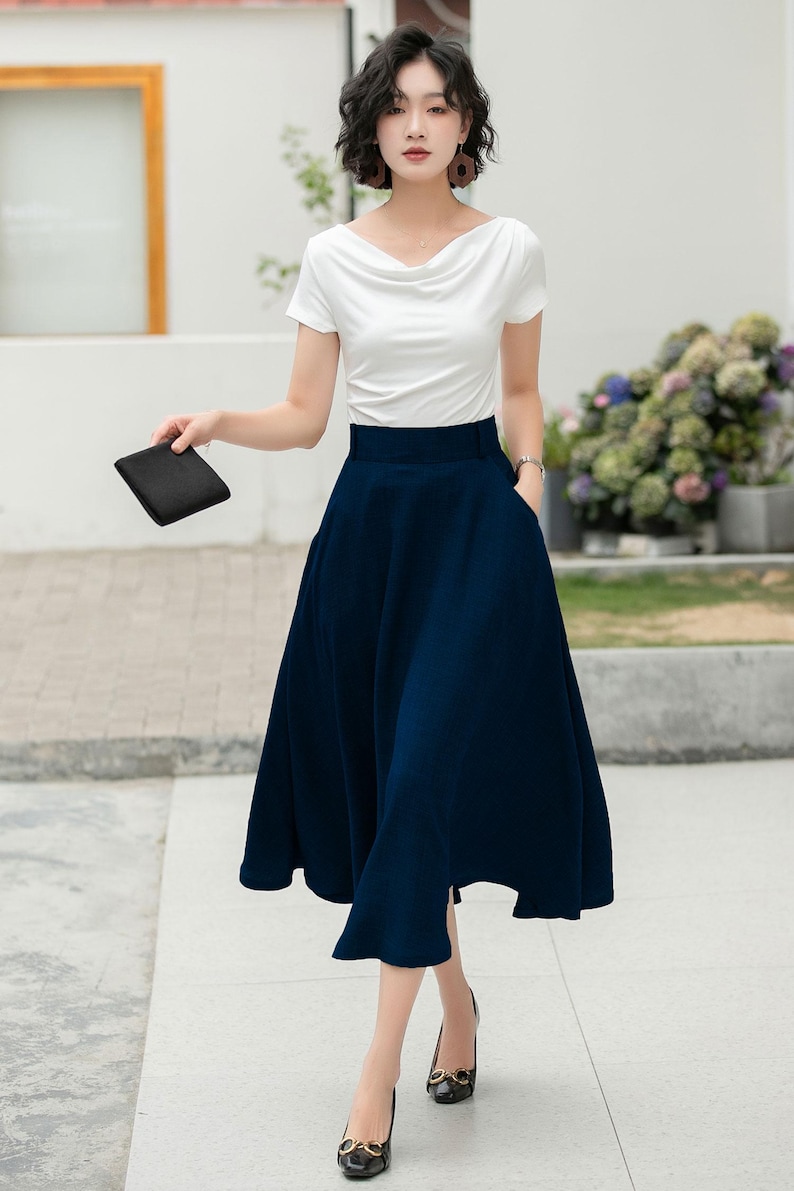 May include: A woman wearing a white short-sleeved top and a dark blue midi skirt with a high waist. She is wearing black heels with gold accents.