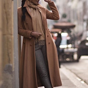 May include: A woman wearing a brown fedora, a long brown coat, gray pants, and black loafers. She is standing next to a building and holding her hat in her hand. The text "@the_Angelique_Noire" is at the bottom of the image.