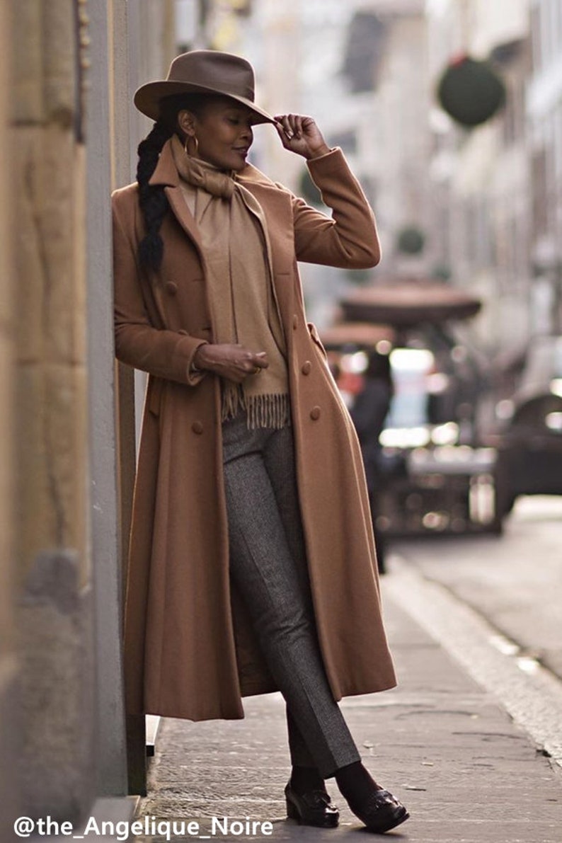 May include: A woman wearing a brown fedora, a long brown coat, gray pants, and black loafers. She is standing next to a building and holding her hat in her hand. The text "@the_Angelique_Noire" is at the bottom of the image.