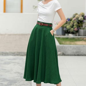 May include: A woman wearing a white t-shirt, a green midi skirt with a brown belt, and a straw hat. She is standing on a sidewalk.
