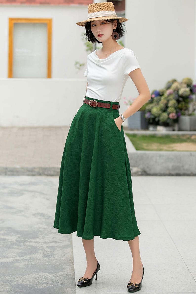 May include: A woman wearing a white t-shirt, a green midi skirt with a brown belt, and a straw hat. She is standing on a sidewalk.