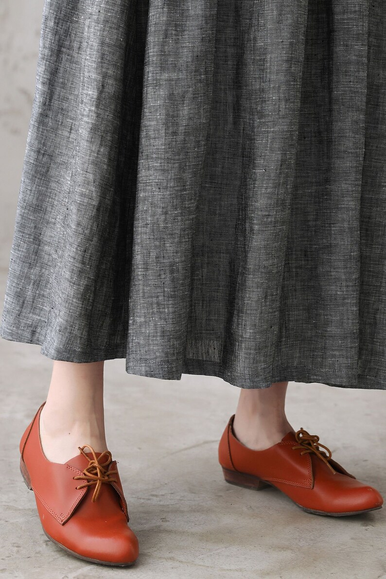 May include: A pair of brown leather lace-up shoes with a low heel. The shoes are worn by a person wearing a gray skirt.