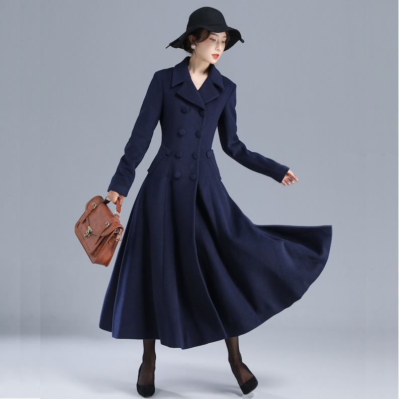 May include: A navy blue double-breasted wool coat with a flared skirt and a classic collar. The coat has a fitted bodice and a long, flowing skirt. The model is wearing a black hat and holding a brown leather bag.