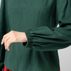 May include: A dark green long-sleeved shirt with gathered cuffs. The shirt has a fitted bodice and a relaxed fit through the sleeves.