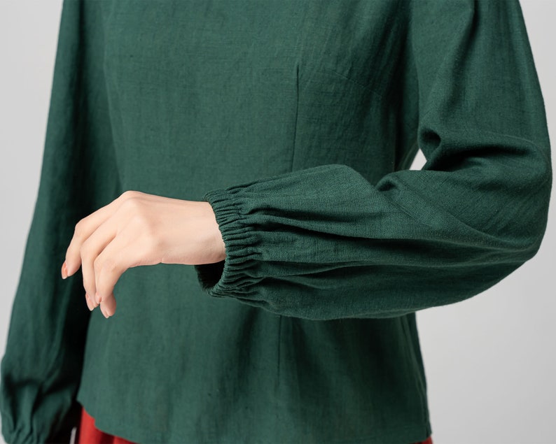 May include: A dark green long-sleeved shirt with gathered cuffs. The shirt has a fitted bodice and a relaxed fit through the sleeves.