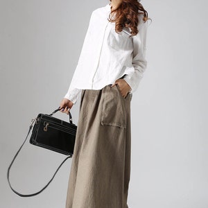 May include: A woman wearing a white long-sleeved shirt and a long, brown, linen skirt with a large pocket. She is carrying a black handbag with a shoulder strap and is wearing brown ankle boots.