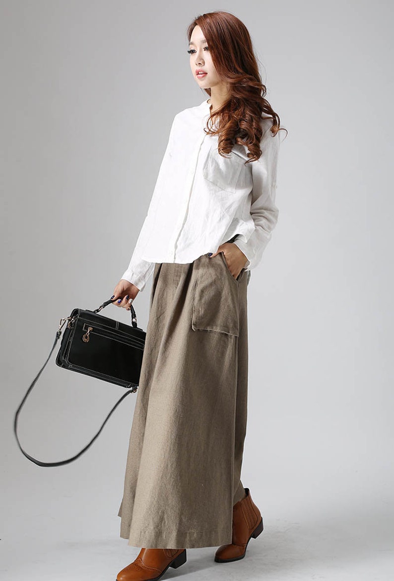 May include: A woman wearing a white long-sleeved shirt and a long, brown, linen skirt with a large pocket. She is carrying a black handbag with a shoulder strap and is wearing brown ankle boots.