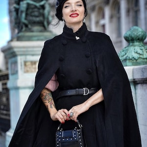 May include: A woman wearing a black cape with a black hat and a black purse with silver studs. She is standing in front of a stone building.