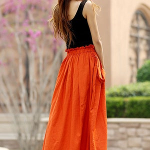 May include: A woman's outfit featuring a black tank top and a long, vibrant orange skirt. The skirt has a gathered waist and falls to mid-calf. The outfit is paired with gold sandals. The background includes greenery and a building.