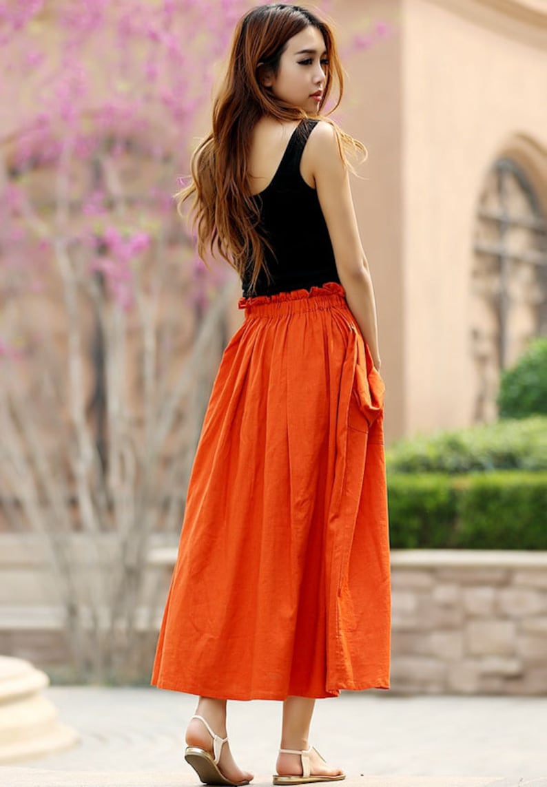 May include: A woman's outfit featuring a black tank top and a long, vibrant orange skirt. The skirt has a gathered waist and falls to mid-calf. The outfit is paired with gold sandals. The background includes greenery and a building.