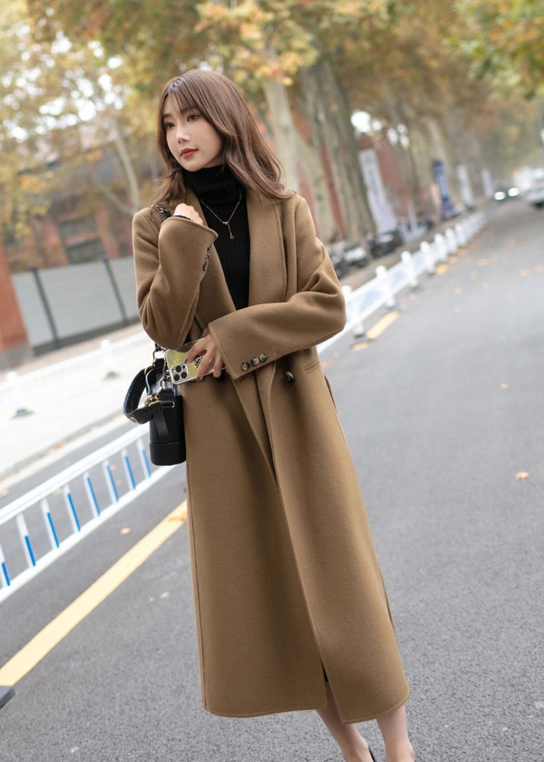 May include: A brown long-sleeved coat with a double-breasted closure and a belt. The coat has a collar and is worn by a person with long brown hair.