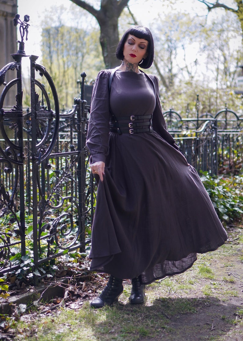May include: A woman wearing a long, dark brown dress with a black leather corset belt. She is standing in front of a black metal fence with green foliage.