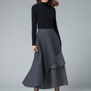 May include: A woman wearing a black turtleneck sweater and a gray layered skirt. The skirt has a longer, flowing layer on top of a shorter, patterned layer.