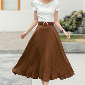 May include: A woman wearing a white short-sleeved shirt, a brown linen skirt with a brown leather belt, and a straw hat. She is standing on a sidewalk.