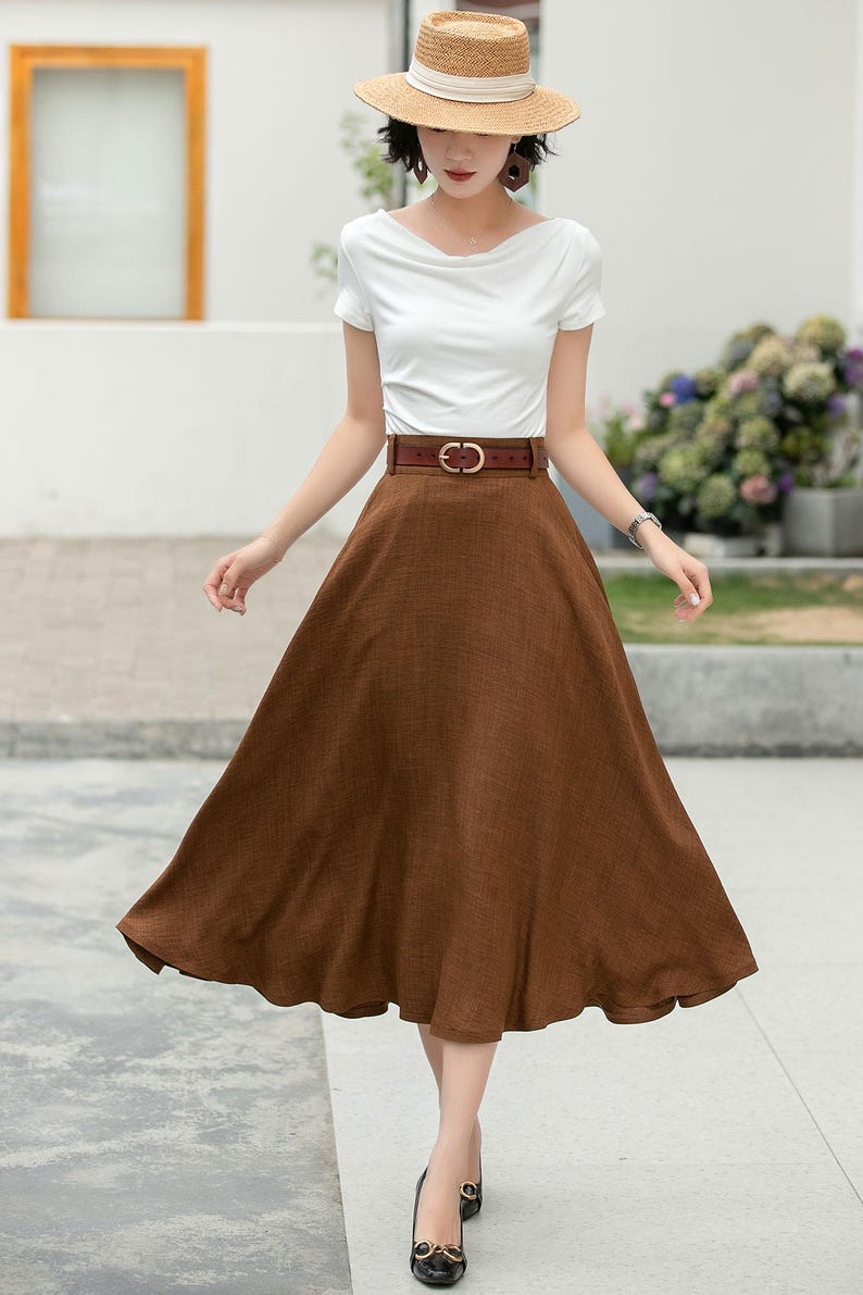 May include: A woman wearing a white short-sleeved shirt, a brown linen skirt with a brown leather belt, and a straw hat. She is standing on a sidewalk.