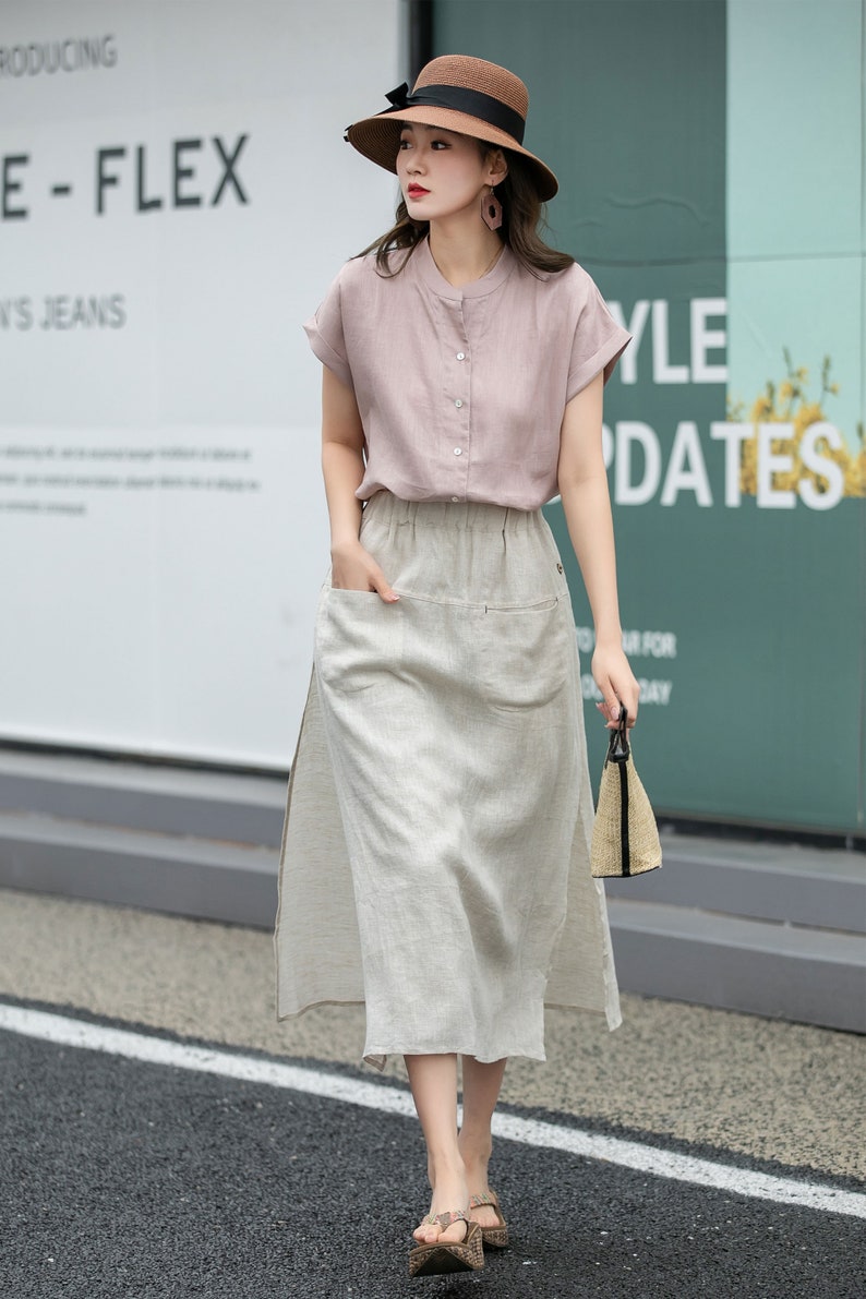 May include: A woman wearing a light pink linen shirt with a button-up collar and short sleeves, a beige linen skirt with a side slit and a large pocket, a brown straw hat with a black ribbon, and brown sandals. She is carrying a small straw bag.