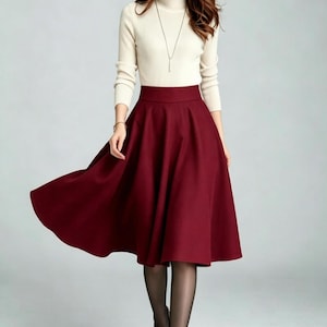 May include: A cream-colored long-sleeve mock neck sweater paired with a solid deep burgundy midi-length skirt. The skirt features a high-waisted design with a flared, A-line silhouette that falls below the knees, complemented by sheer black tights.