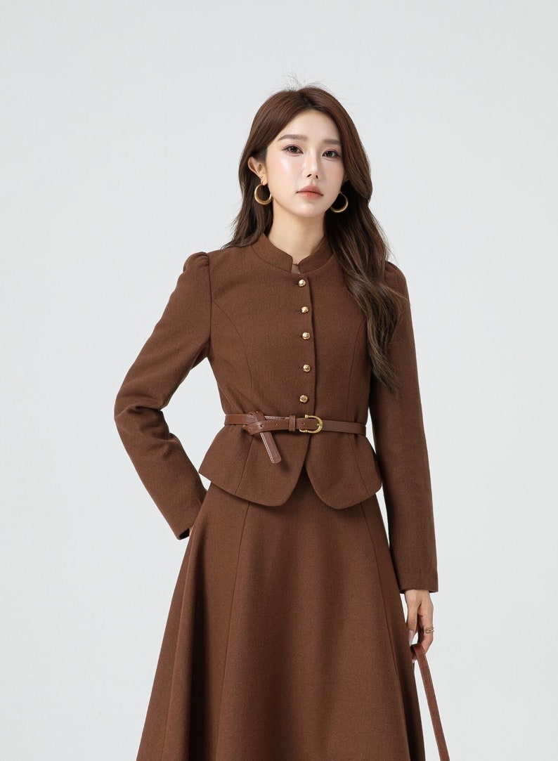 May include: A brown wool skirt suit with a fitted jacket and a flared skirt. The jacket has a mandarin collar, gold buttons, and a brown leather belt.