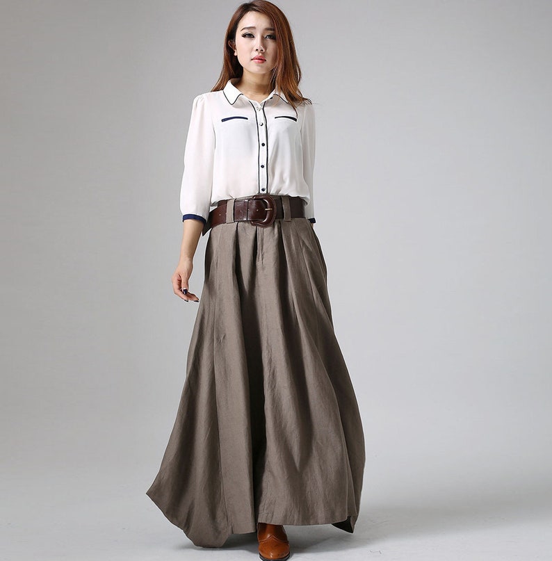 May include: A brown linen maxi skirt with a wide brown leather belt with a large buckle. The skirt has a pleated design and is worn with a white button-down shirt with a navy blue trim.