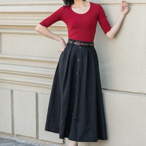 May include: A woman wearing a red short-sleeved shirt and a black button-front skirt with a brown belt. She is standing in front of a white wall.