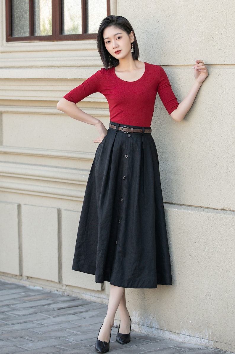 May include: A woman wearing a red short-sleeved shirt and a black button-front skirt with a brown belt. She is standing in front of a white wall.