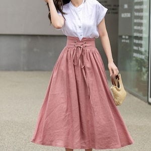 May include: A woman wearing a white short-sleeved shirt with a pink midi skirt with a lace-up detail at the waist. She is holding a straw bag.