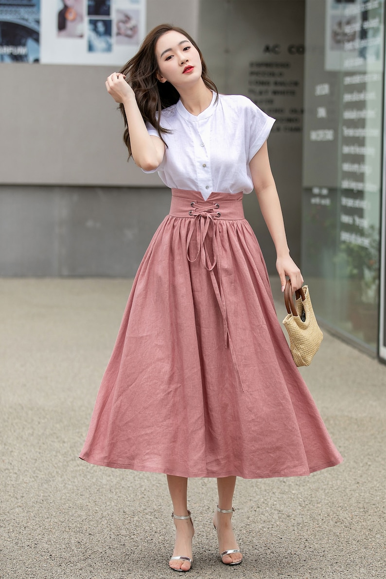 May include: A woman wearing a white short-sleeved shirt with a pink midi skirt with a lace-up detail at the waist. She is holding a straw bag.