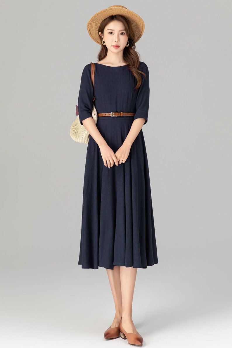 May include: A navy blue midi dress with a brown belt and a straw hat. The dress has a boat neck and 3/4 sleeves.