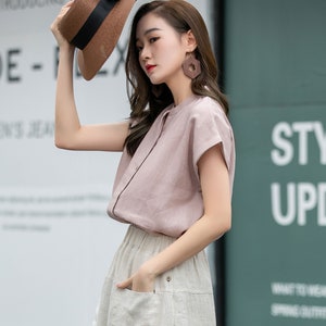 May include: A woman wearing a light pink linen blouse with a white linen skirt. She is holding a brown straw hat with a black ribbon and bow. The hat has a wide brim. She is wearing large brown hexagonal earrings.
